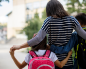 A parent is walking with two children, one on each side, down a pathway. The parent is wearing a striped shirt and has their arms around both children. The child on the left is carrying a pink backpack, and the child on the right is carrying a black and yellow backpack. The background shows a blurred view of buildings and trees, suggesting they are on their way to school.