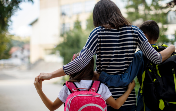 A parent is walking with two children, one on each side, down a pathway. The parent is wearing a striped shirt and has their arms around both children. The child on the left is carrying a pink backpack, and the child on the right is carrying a black and yellow backpack. The background shows a blurred view of buildings and trees, suggesting they are on their way to school.