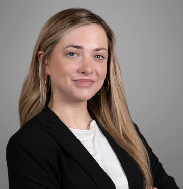 Courtney Gardner, a family law paralegal in Winston-Salem, NC, poses confidently for a professional headshot. She has long, straight blonde hair and is wearing a black blazer over a white top, complemented by gold hoop earrings. She smiles subtly and stands against a plain gray background.