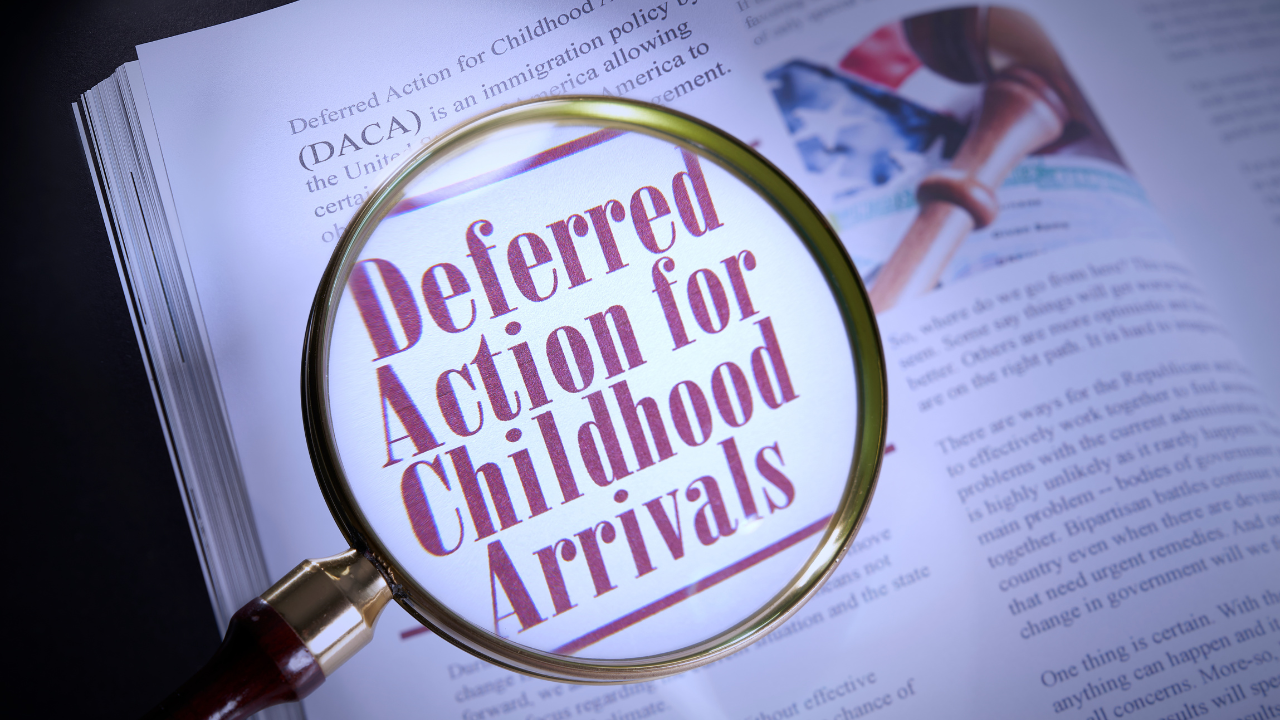 A magnifying glass focusing on the words "Deferred Action for Childhood Arrivals" printed in red, bold letters on a book page. The surrounding text, slightly blurred, includes a definition of DACA. A gavel and an American flag are visible in the background on the same page.