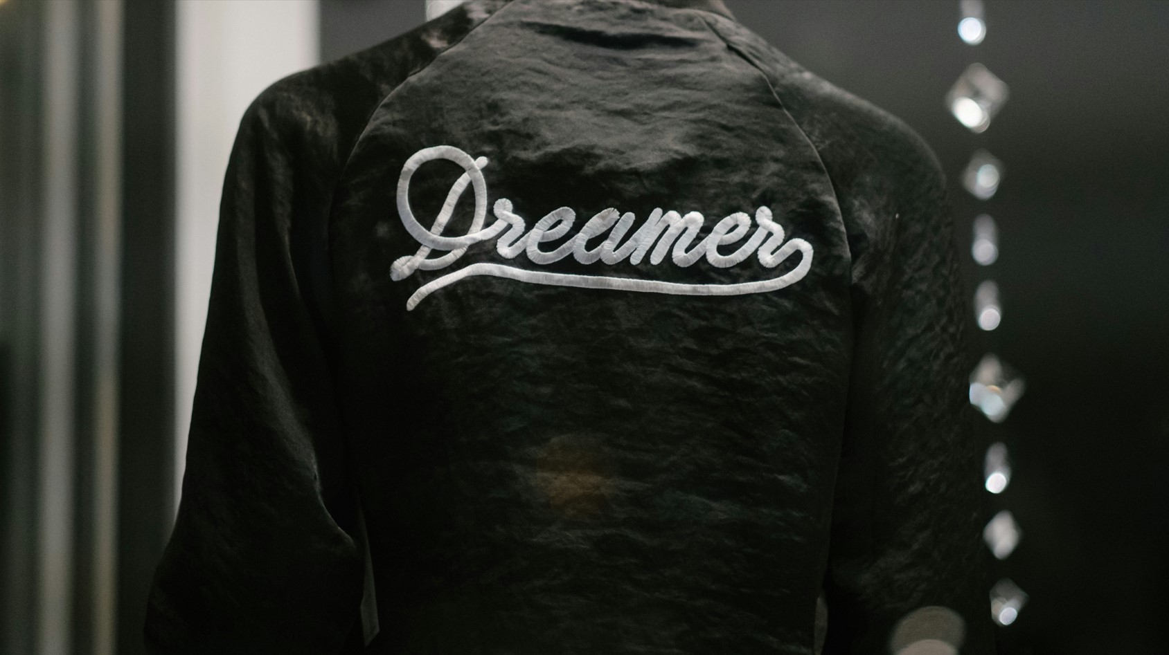 The image shows the back of a person wearing a black jacket with the word "Dreamer" written in white, cursive script across the upper back in reference to DACA. The setting appears to be indoors, with some decorative hanging elements visible in the blurred background.