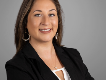 Heather Wall, a family law paralegal, poses in a professional headshot. She has long, brown hair and is wearing a black blazer over a white top, accessorized with hoop earrings and a necklace. Heather is smiling confidently against a neutral gray background.