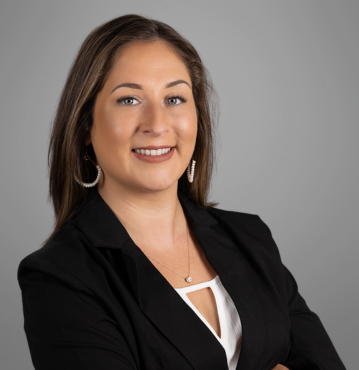 Heather Wall, a family law paralegal, poses in a professional headshot. She has long, brown hair and is wearing a black blazer over a white top, accessorized with hoop earrings and a necklace. Heather is smiling confidently against a neutral gray background.