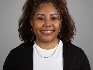 This image is a professional headshot of Christa Anderson, who works in Client Relations for Dummit Fradin Attorneys at Law. She is smiling, has curly hair, and is wearing a white top with a black cardigan. She is also wearing a gold necklace and hoop earrings. The background is a plain, neutral grey.