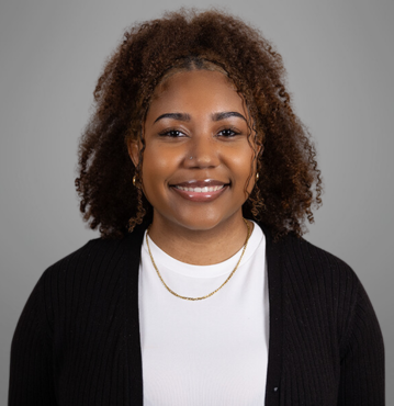 This image is a professional headshot of Christa Anderson, who works in Client Relations for Dummit Fradin Attorneys at Law. She is smiling, has curly hair, and is wearing a white top with a black cardigan. She is also wearing a gold necklace and hoop earrings. The background is a plain, neutral grey.