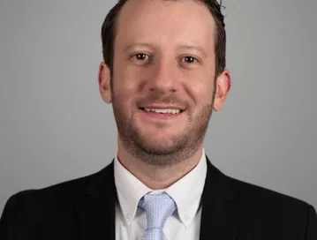 Thomas Mahon, a criminal defense attorney in Greensboro, NC, facing forward, wearing a black suit jacket, white shirt, and light blue tie, smiling.