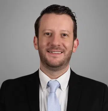 Thomas Mahon, a criminal defense attorney in Greensboro, NC, facing forward, wearing a black suit jacket, white shirt, and light blue tie, smiling.